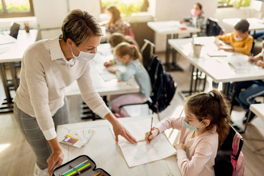 Eine Lehrerin trägt eine Gesichtsmaske, während sie eine Schülerin während eines Unterrichts im Klassenzimmer unterstützt (Symbolbild: iStock/Drazen Zigic)