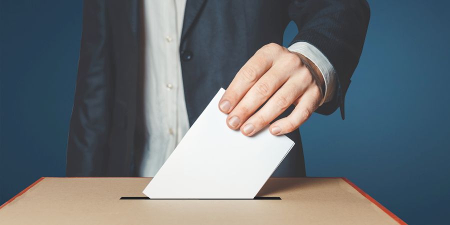 Ein Mann im Anzug steckt einen Umschlag in eine Wahlurne (Symbolbild: iStock/Sergey Tinyakov)