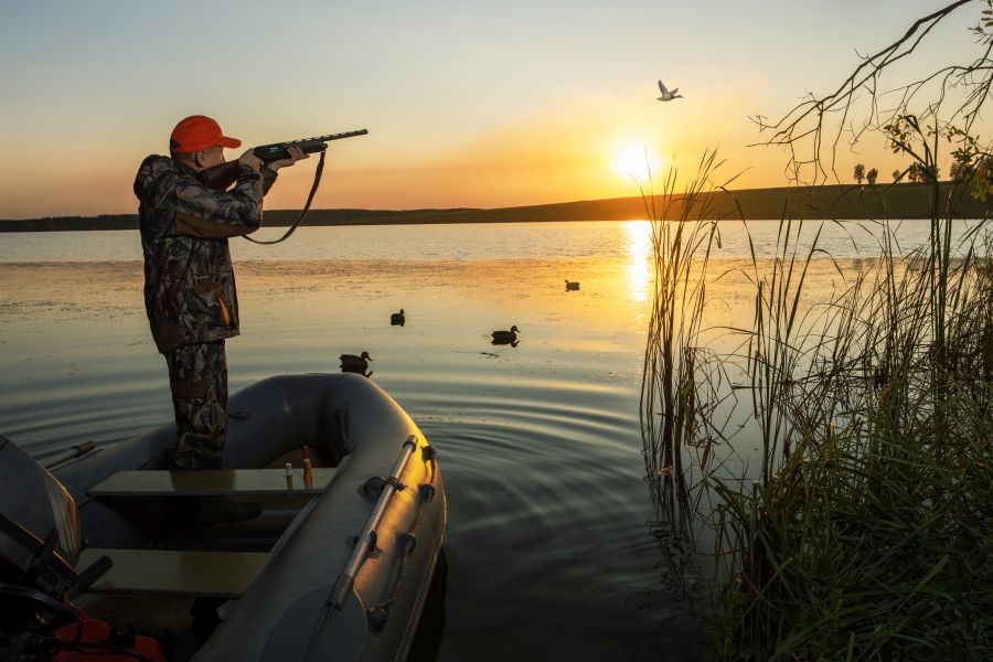 Ein Jäger, der in einem Boot auf einem See steht, zielt mit einer Selbstladeflinte auf eine vorbeifliegende Ente. (Symbolbild: iStock/Sergei Chuyko)