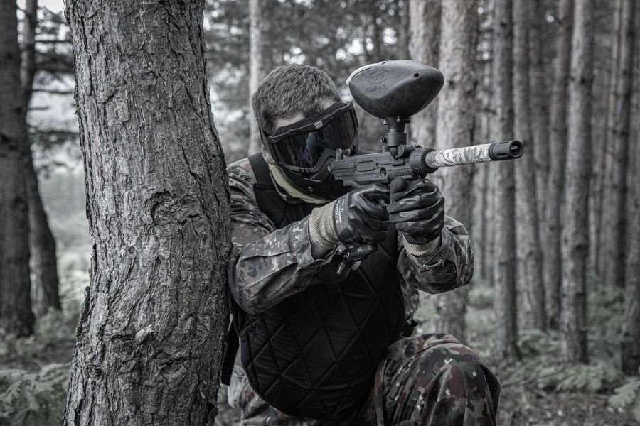 Ein Mann in Tarnkleidung und Schutzausrüstung, der kniend in einem Wald einen Paintball-Markierer im Anschlag hält. (Symbolbild: mnba auf Pixabay)