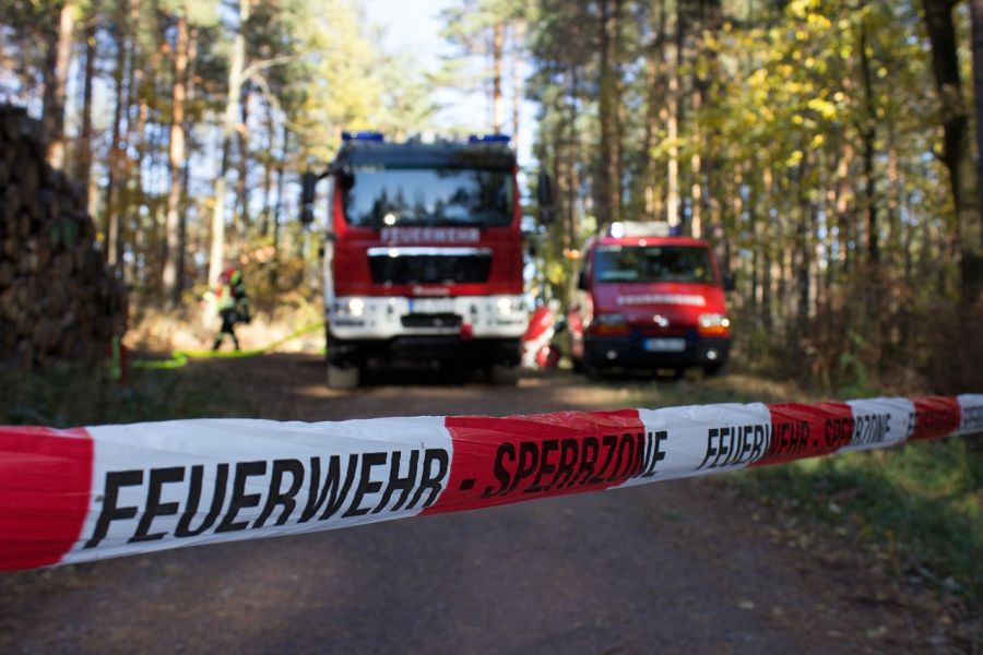 Feuerwehrfahrzeuge und Einsatzkräfte in einem Waldstück. Im Vordergund ist ein Absperrband mit der Aufschrift „Feuerwehr – Sperrzone“ über einen Waldweg zu sehen. (Symbolbild: Ronny auf Pixabay)